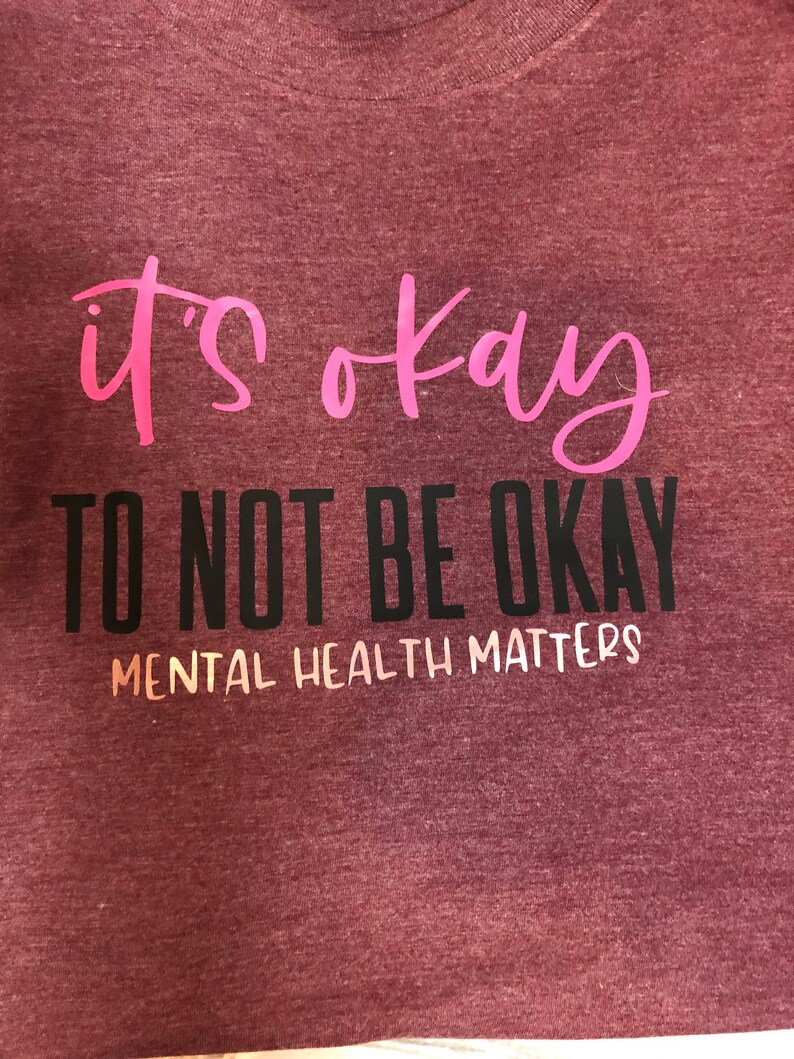 May include: A maroon t-shirt with the words "it's okay" in pink script. Below, in black block letters, it reads "TO NOT BE OKAY". The phrase "MENTAL HEALTH MATTERS" is printed in white beneath the black text.