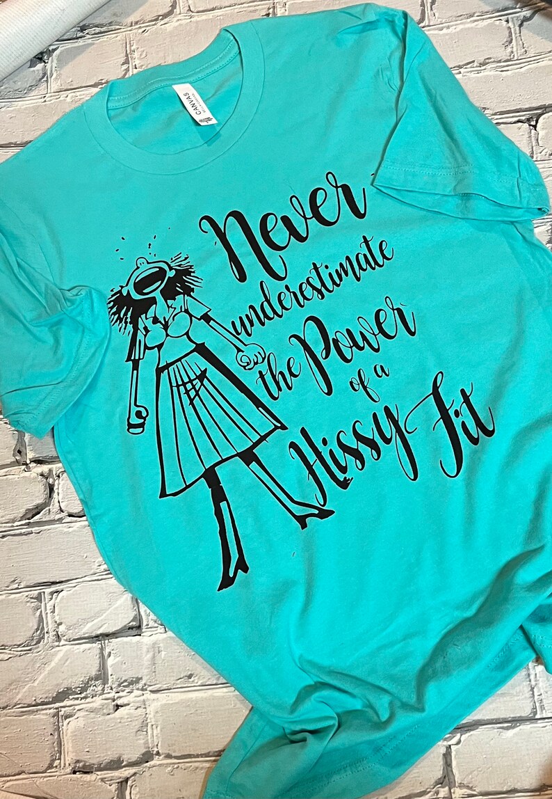 Puede incluir: Una camiseta turquesa con un gr&aacute;fico negro de una mujer con una expresi&oacute;n dram&aacute;tica y el texto "Nunca subestimes el poder de una rabieta."