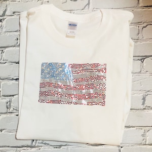 May include: White t-shirt with a red, white, and blue American flag design made of rhinestones.