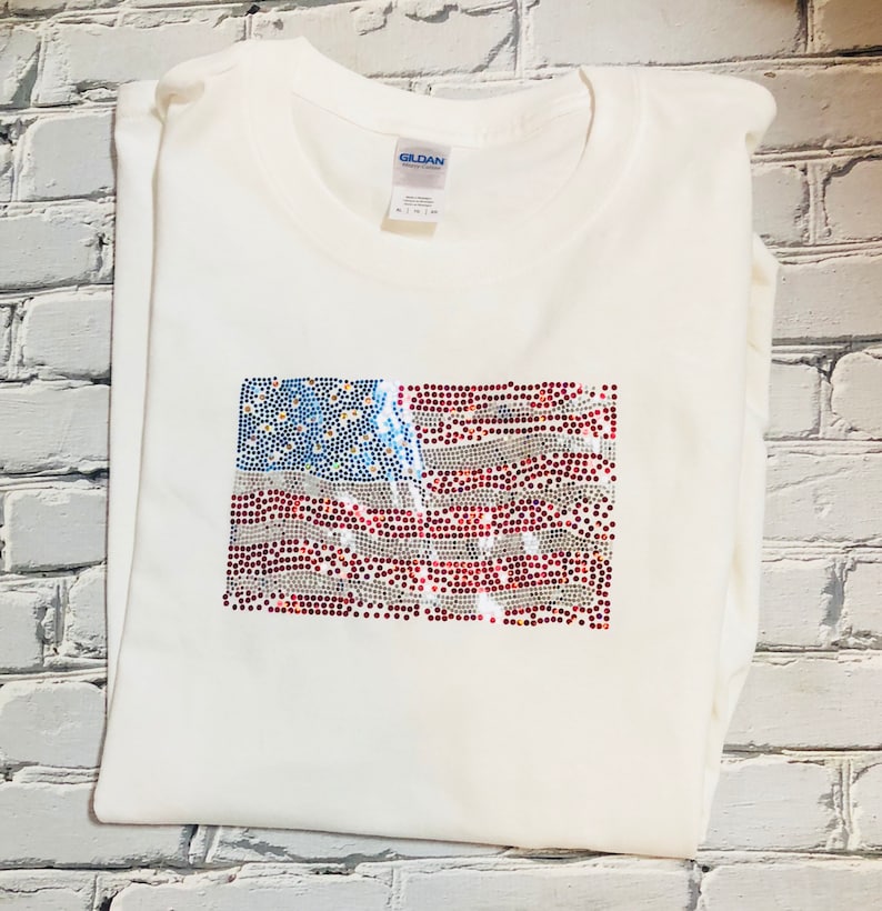 May include: White t-shirt with a red, white, and blue American flag design made of rhinestones.