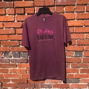 May include: Maroon t-shirt with the text "it's okay TO NOT BE OKAY MENTAL HEALTH MATTERS" in pink and black. The shirt is hanging on a black hanger against a red brick wall.