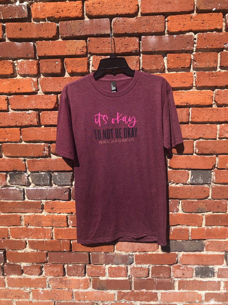 May include: Maroon t-shirt with the text "it's okay TO NOT BE OKAY MENTAL HEALTH MATTERS" in pink and black. The shirt is hanging on a black hanger against a red brick wall.