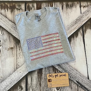 May include: Gray t-shirt with a sparkly American flag design. The flag is made of rhinestones in red, white, and blue. The shirt is folded in half and the bottom of the shirt is out of frame. The shirt is on a wooden background. The text "let's get social" is on a wooden sign below the shirt. The text "Designs by Millie" is on the sign below the text "let's get social". The text "#designs_by_millie" is on the sign below the text "Designs by Millie".