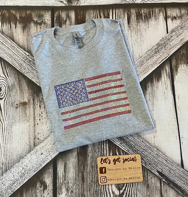 May include: Gray t-shirt with a sparkly American flag design. The flag is made of rhinestones in red, white, and blue. The shirt is folded in half and the bottom of the shirt is out of frame. The shirt is on a wooden background. The text "let's get social" is on a wooden sign below the shirt. The text "Designs by Millie" is on the sign below the text "let's get social". The text "#designs_by_millie" is on the sign below the text "Designs by Millie".