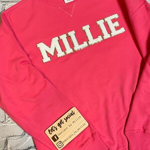 Puede incluir: Sudadera rosa con el nombre "MILLIE" en letras blancas con detalles de purpurina dorada. La sudadera tiene una etiqueta de madera adjunta que dice "let's get social" con iconos de redes sociales y el texto "Designs by Millie" y "@designs_by_millie".