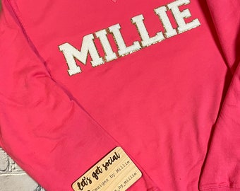May include: Pink sweatshirt with the name "MILLIE" in white letters with gold glitter accents. The sweatshirt has a wooden tag attached that says "let's get social" with social media icons and the text "Designs by Millie" and "@designs_by_millie".