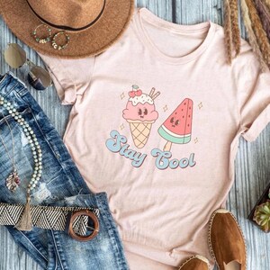 Puede incluir: Una camiseta rosa con un gr&aacute;fico de un cono de helado y una paleta de sand&iacute;a con el texto "Stay Cool".