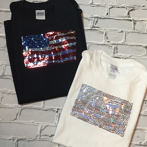 May include: Two t-shirts with an American flag design made of iridescent sequins. One shirt is black and the other is white.