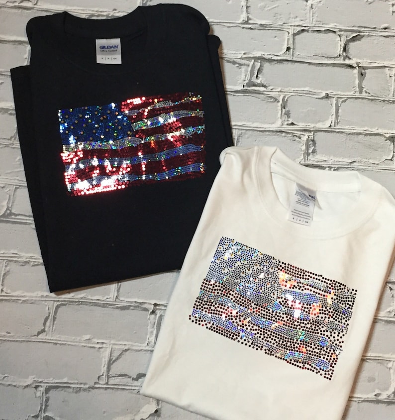 May include: Two t-shirts with an American flag design made of iridescent sequins. One shirt is black and the other is white.