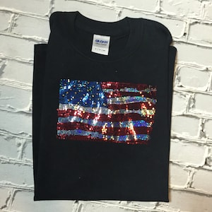 May include: Black t-shirt with a sequined American flag design. The flag is red, white, and blue with iridescent sequins.
