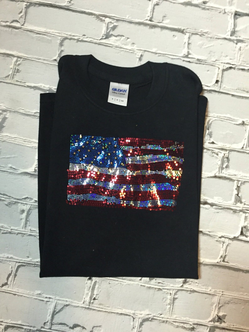 May include: Black t-shirt with a sequined American flag design. The flag is red, white, and blue with iridescent sequins.