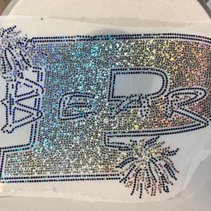 May include: A rhinestone applique spelling "PURS" in a decorative font, with a shimmering, iridescent finish. The design incorporates embellishments and is set on a white fabric backing.