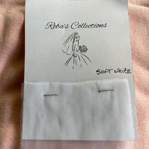 May include: A close-up of a white tulle fabric sample with two pins holding it in place. The fabric is labeled "Soft White" and is displayed on a white card with a drawing of a bride in a wedding dress. The card also has the text "Reba's Collections" printed on it.