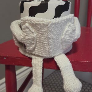 Op de afbeelding: Een handgemaakte, witte gebreide pop met een zwart-wit patroon op het hoofd, zittend op een rode stoel. De pop heeft armen, benen en een lichaam, allemaal gemaakt van wit garen. De tekst "knit pattern pdf" staat bovenaan.