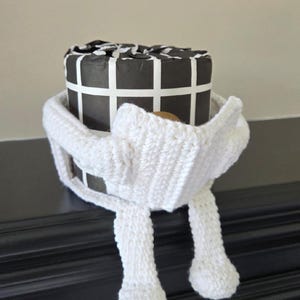 Op de afbeelding: Een witte gehaakte sjaal wikkelt zich om een zwart-witte geschenkdoos. De woorden "crochet pattern" staan bovenaan de afbeelding. De tekst "Carol Hladik Designs" staat onderaan de afbeelding.