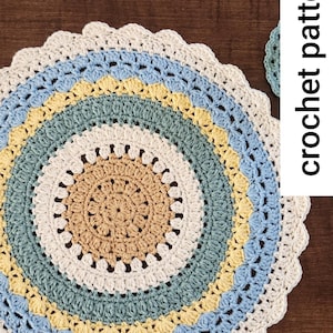 May include: A crocheted placemat with a multi-coloured design. The placemat is made with a variety of shades of blue, green, yellow, and beige yarn. The placemat has a scalloped edge.