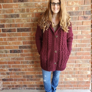 May include: A maroon crocheted cardigan with a cable knit pattern and button closure. The cardigan has a hood and pockets. The model is wearing the cardigan with blue jeans and brown boots.
