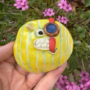 Pode incluir: Uma escultura de cerâmica artesanal e caprichosa de uma criatura amarela listrada com um único olho, um sorriso dentuço e uma tira vermelha. A escultura tem um olho azul e um pequeno chifre. O fundo apresenta flores cor-de-rosa e folhagem verde.