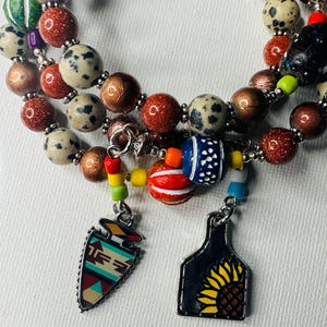 May include: A multi-strand bracelet with brown, white, and black beads. The bracelet has two charms: a silver arrowhead with a Native American design and a silver tag with a sunflower.