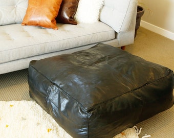 May include: A black leather pouffe ottoman with a square top and a tufted design. The pouffe is sitting on a white rug with a light brown sofa in the background.