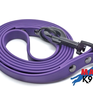 Może przedstawiać: Fioletowa gumowa smycz dla psa z czarnym metalowym karabińczykiem i dwoma czarnymi metalowymi kółkami D. Smycz jest zwinięta i ma napis "MA K9" na końcu.