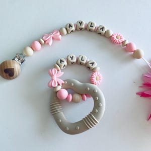 May include: A baby pacifier clip and teether set in soft pink, beige, and gray. The pacifier clip features the name "AYLA" spelled out in letter beads, a bow, and a wooden heart clip. The teether is heart-shaped with the name "AYLA" and a flower.