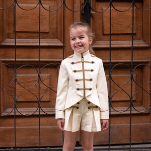 Può includere: Una bambina che indossa una giacca bianca con bottoni dorati e pantaloncini abbinati. Sta sorridendo e in piedi davanti a una porta di legno con un cancello di metallo nero.