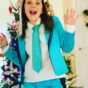 Pode incluir: Uma pessoa jovem vestindo um terno azul turquesa com uma camisa branca e uma gravata azul turquesa. A pessoa est&aacute; sorrindo e tem os bra&ccedil;os levantados no ar. H&aacute; uma &aacute;rvore de Natal no fundo.