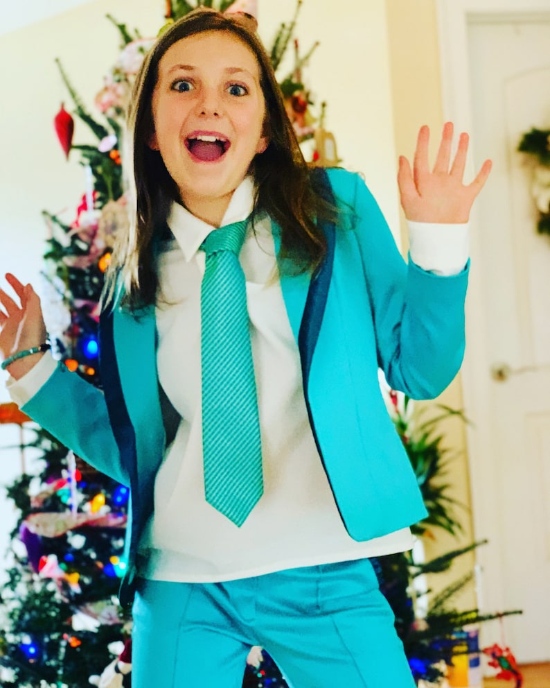 Pode incluir: Uma pessoa jovem vestindo um terno azul turquesa com uma camisa branca e uma gravata azul turquesa. A pessoa est&aacute; sorrindo e tem os bra&ccedil;os levantados no ar. H&aacute; uma &aacute;rvore de Natal no fundo.