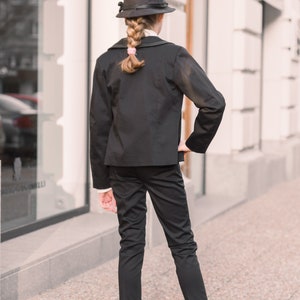 Pode incluir: Uma pessoa jovem vestindo um terno preto com um chap&eacute;u preto e sapatos pretos. A pessoa est&aacute; em p&eacute; na frente de um pr&eacute;dio com uma grande janela.
