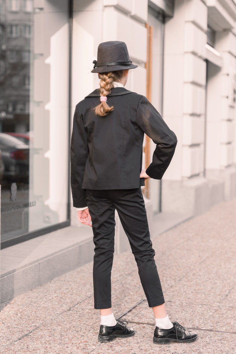 Pode incluir: Uma pessoa jovem vestindo um terno preto com um chap&eacute;u preto e sapatos pretos. A pessoa est&aacute; em p&eacute; na frente de um pr&eacute;dio com uma grande janela.