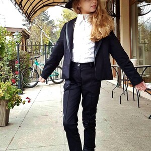 Pode incluir: Uma pessoa jovem vestindo um blazer preto, uma camisa branca de colarinho, cal&ccedil;as pretas e botas pretas com solas cor-de-rosa. Ela est&aacute; usando um chap&eacute;u fedora preto e tem cabelo loiro comprido.
