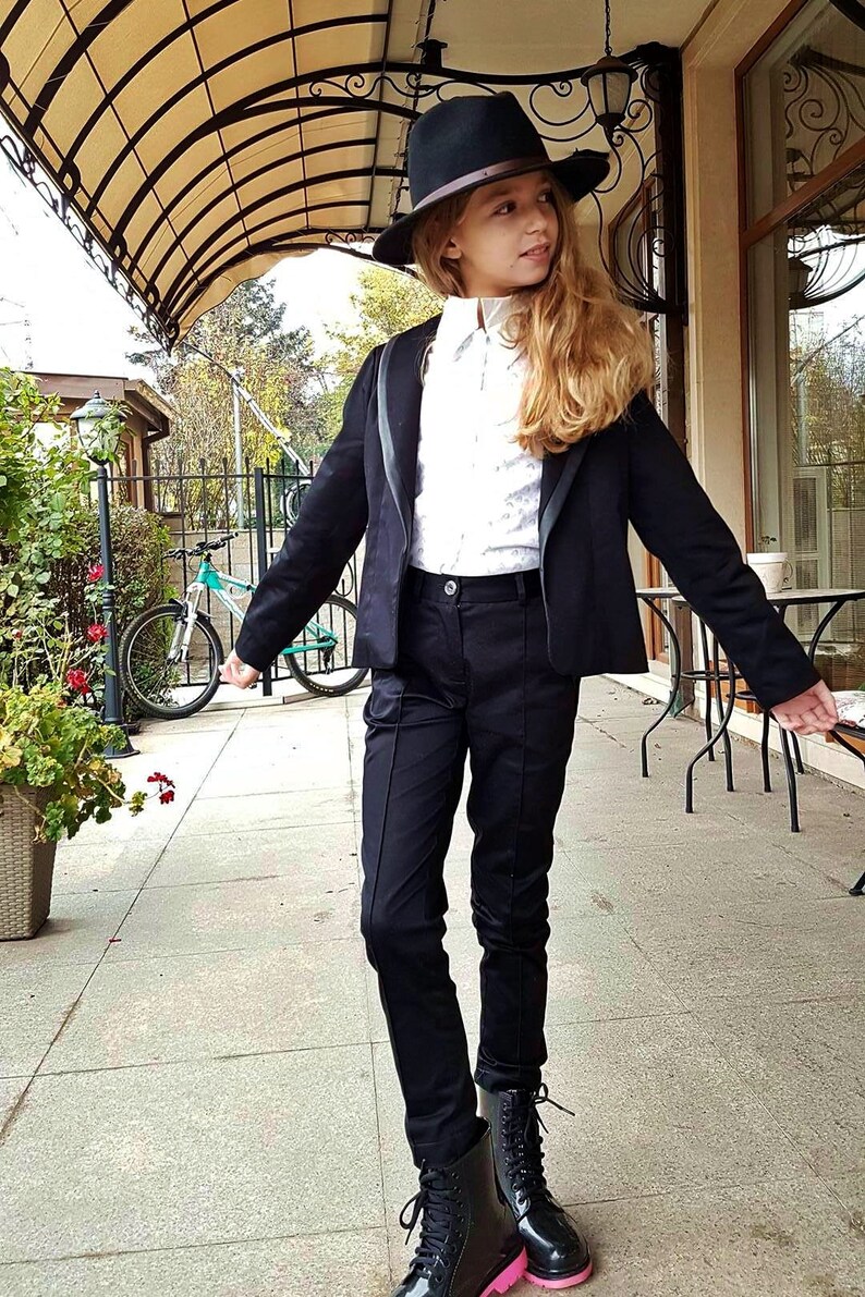 Pode incluir: Uma pessoa jovem vestindo um blazer preto, uma camisa branca de colarinho, cal&ccedil;as pretas e botas pretas com solas cor-de-rosa. Ela est&aacute; usando um chap&eacute;u fedora preto e tem cabelo loiro comprido.