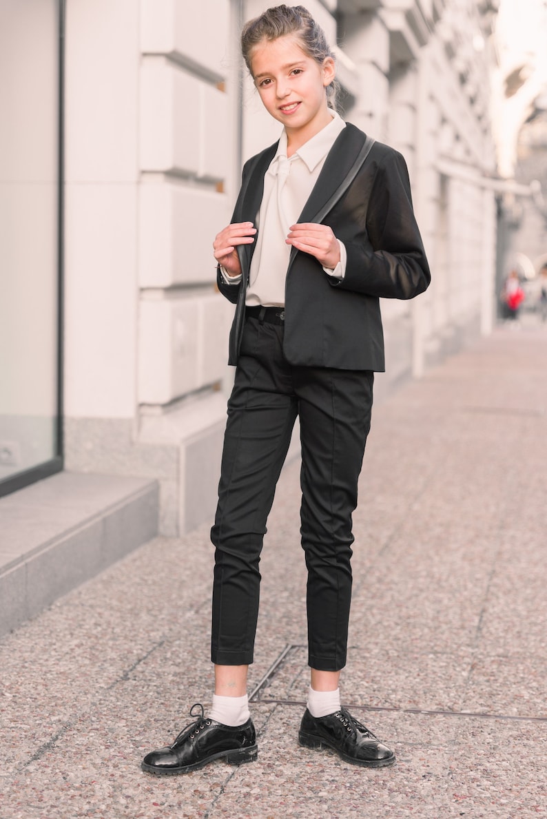 Pode incluir: Uma pessoa jovem vestindo um blazer preto com uma camisa branca e cal&ccedil;as pretas. Ela est&aacute; em p&eacute; em uma cal&ccedil;ada na frente de um pr&eacute;dio.