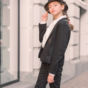 Pode incluir: Uma pessoa jovem vestindo uma jaqueta de terno preta e cal&ccedil;as pretas com uma camisa branca de colarinho por baixo. Ela est&aacute; usando um chap&eacute;u fedora preto e tem o cabelo puxado para tr&aacute;s em uma tran&ccedil;a. A pessoa est&aacute; em p&eacute; na frente de um pr&eacute;dio com uma fachada branca.