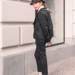 Pode incluir: Uma pessoa jovem vestindo um terno preto com um chap&eacute;u fedora preto. A pessoa est&aacute; em p&eacute; em frente a uma parede branca.