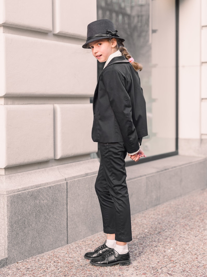 Pode incluir: Uma pessoa jovem vestindo um terno preto com um chap&eacute;u fedora preto. A pessoa est&aacute; em p&eacute; em frente a uma parede branca.