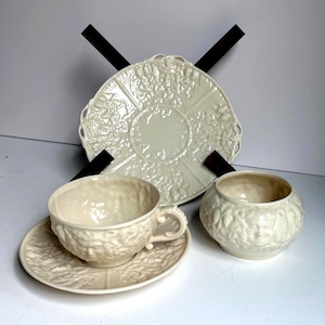 May include: Cream-colored ceramic tea set with embossed floral and face designs. The set includes a teacup and saucer, a sugar bowl, and a large serving plate. The pieces have a textured surface and a delicate, vintage aesthetic.