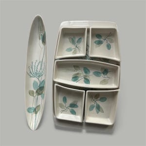 May include: A white ceramic serving set with a long oval platter and a rectangular tray with six smaller dishes. Each piece is decorated with a teal and gray leaf design. The set is ideal for serving appetizers or snacks.