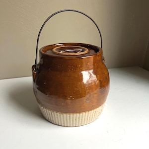 May include: A vintage brown stoneware crock with a lid and a metal handle. The crock has a glossy finish and a light beige, ribbed base. The handle arches over the top, and the lid has a recessed handle. A classic kitchen or farmhouse decor item.