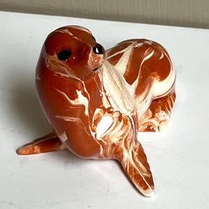 May include: A ceramic seal figurine in shades of brown and white. The seal is posed with its head turned, featuring black eyes and a marbled pattern throughout its body. The figurine is a decorative item.