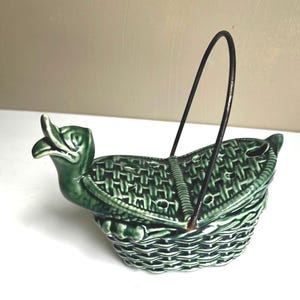 May include: Green ceramic duck-shaped basket with a woven texture and a black metal handle. The duck's head forms the front of the basket, with the beak open. The basket is a decorative item.