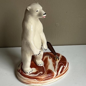 May include: A ceramic polar bear figurine with a pen holder. The bear is white with black accents and a red mouth. The base is oval-shaped with a brown and white marbled design. The pen holder is brown with a gold-colored pen.