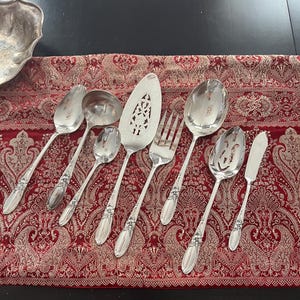 May include: A collection of antique silver serving utensils, including spoons, a ladle, a cake server, a fork, and a butter knife. The silverware features ornate floral detailing on the handles and is arranged on a red paisley patterned tablecloth.