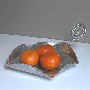 May include: A decorative silver-colored metal serving dish with a leaf-like shape, holding three bright orange tangerines. The dish has a textured, floral design and a curled handle. The tangerines are ripe and fresh.