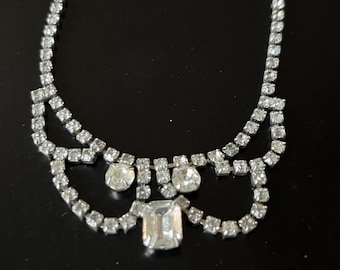 May include: A silver-toned necklace with clear, sparkling rhinestones. The necklace features a series of small stones forming a delicate chain, with larger, round and rectangular stones as focal points. The design includes a curved, layered pattern.