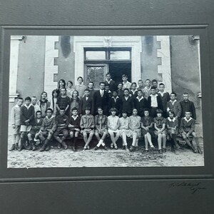 May include: Black and white photograph of a group of children and adults posing in front of a building. The subjects are arranged in rows, some sitting and some standing. The photograph is mounted in a dark gray frame.