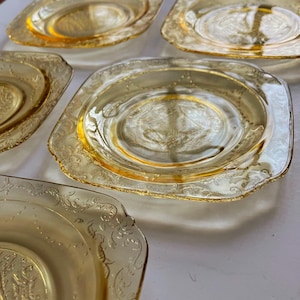 May include: A collection of six square, amber-colored glass plates. Each plate features a decorative embossed floral pattern around the edges and in the center. The plates have a slightly raised rim and a clear, transparent appearance.