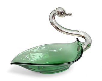 May include: A green glass swan-shaped dish with a clear glass neck and head. The dish has a leaf-like shape with a black rim. The swan's neck curves gracefully, and the head is detailed. The dish is likely for decorative purposes.