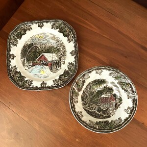May include: A square ceramic plate and a round ceramic bowl, both featuring white centers with detailed winter landscape illustrations of a covered bridge and a mill, surrounded by dark green leaf-patterned borders on the scalloped edges.